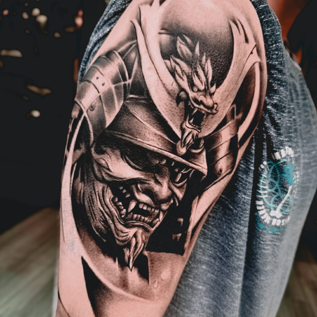 A detailed tattoo on an upper arm shows a fierce samurai wearing a helmet adorned with a dragon motif, with intense facial expression and intricate shading, on a person wearing a sleeveless vest. | CB Ink Tattoo