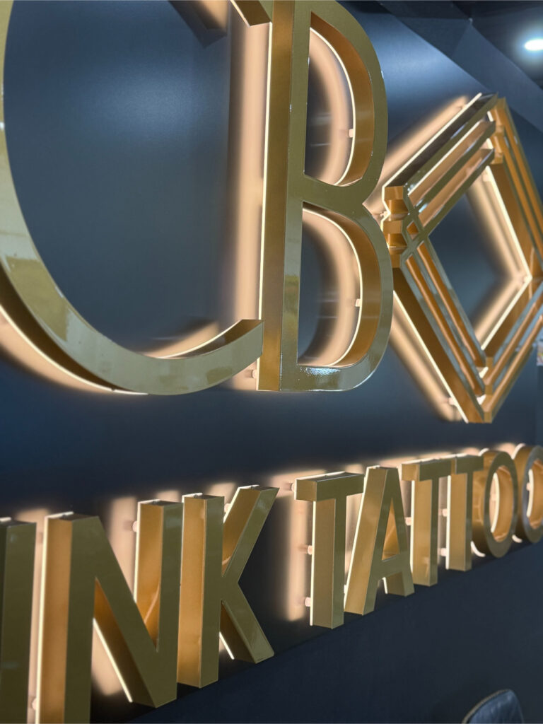 Gold 3D letters and geometric shapes mounted on a dark wall, spelling out part of INK TATTOO. The sign appears to be part of a tattoo studio’s branding or logo. | CB Ink Tattoo