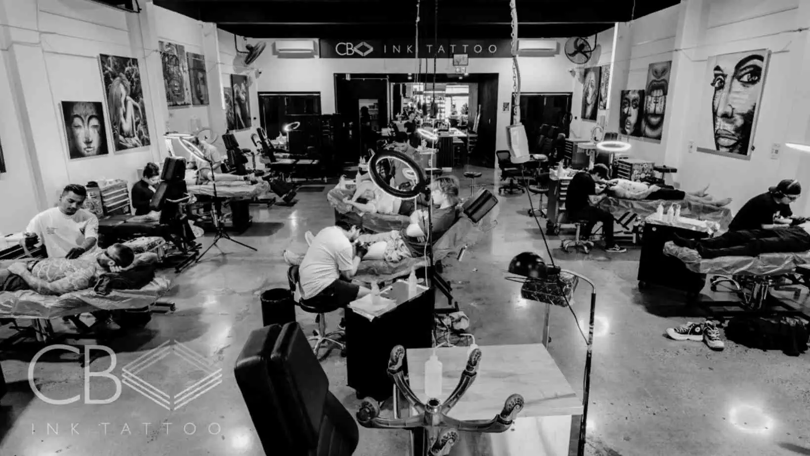 A black-and-white photo of a busy tattoo studio with multiple artists working on clients at various stations. Artworks and tattoo equipment are visible throughout the room. The CB INK TATTOO logo is in the foreground. | CB Ink Tattoo