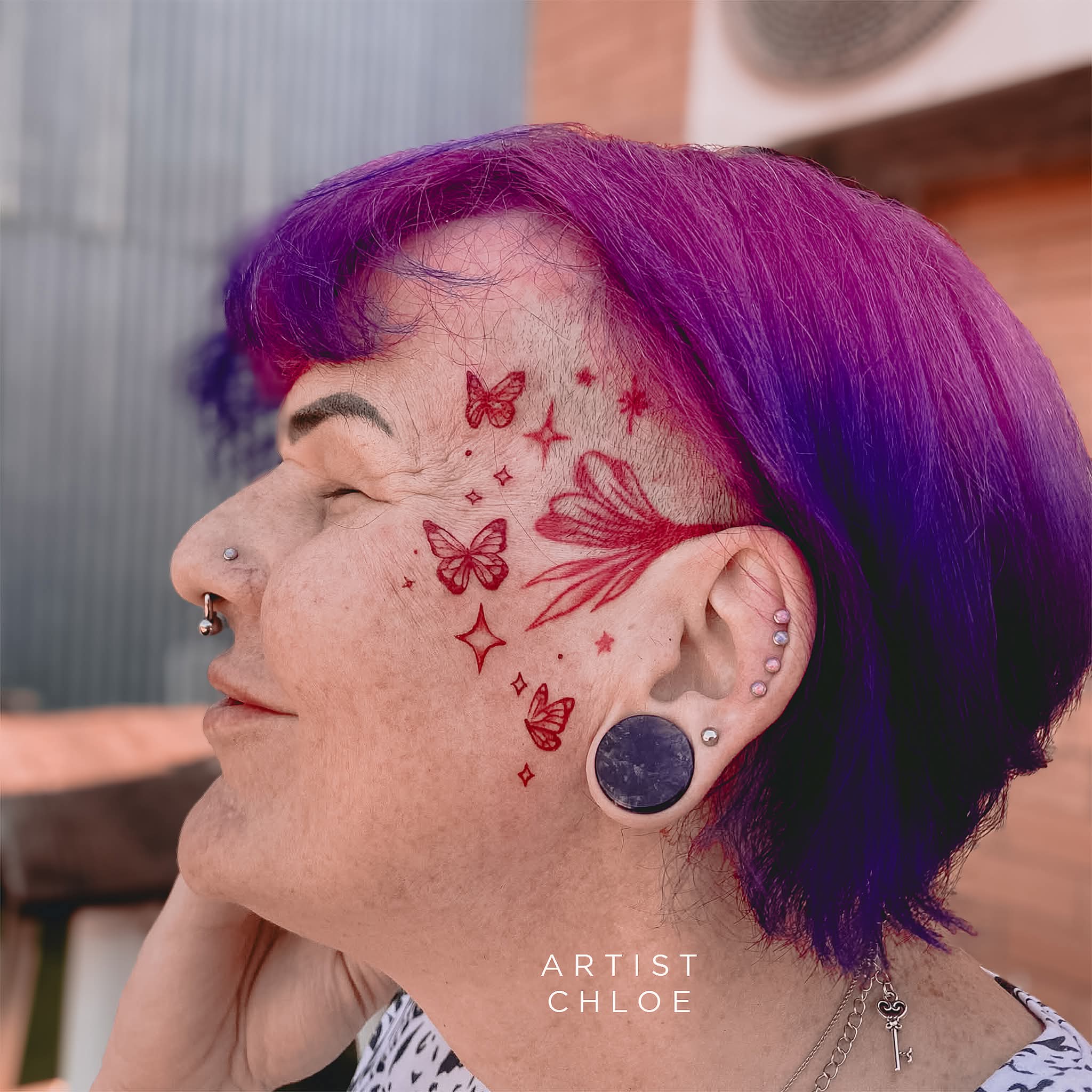 A person with purple hair and multiple ear piercings smiles, showing red tattoos of butterflies, stars, and flowers on their shaven temple—fresh from a recent tattoo touch-up. The text ARTIST CHLOE appears at the bottom. | CB Ink Tattoo
