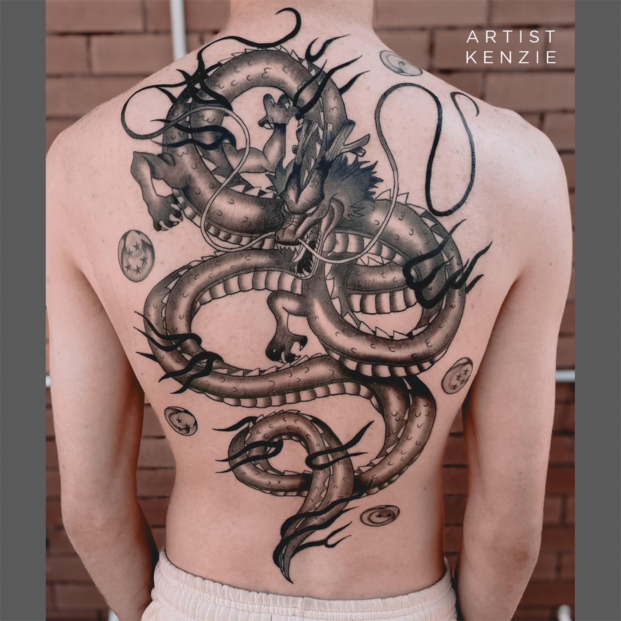 A detailed black and grey tattoo of a large dragon with horns covers a person’s entire back, surrounded by circular symbols. The brick wall background enhances the art. Text in the corner reads, Artist Kenzie—perfect for inspiration before your tattoo touch-up. | CB Ink Tattoo