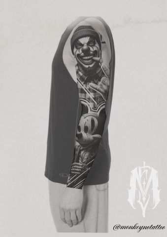 A person wearing a sleeveless black shirt displays a full-sleeve tattoo featuring a sinister clown with a cap and a grinning cartoon character, possibly Mickey Mouse, on their left arm. Tattoo artist logo and Instagram handle are visible. | CB Ink Tattoo